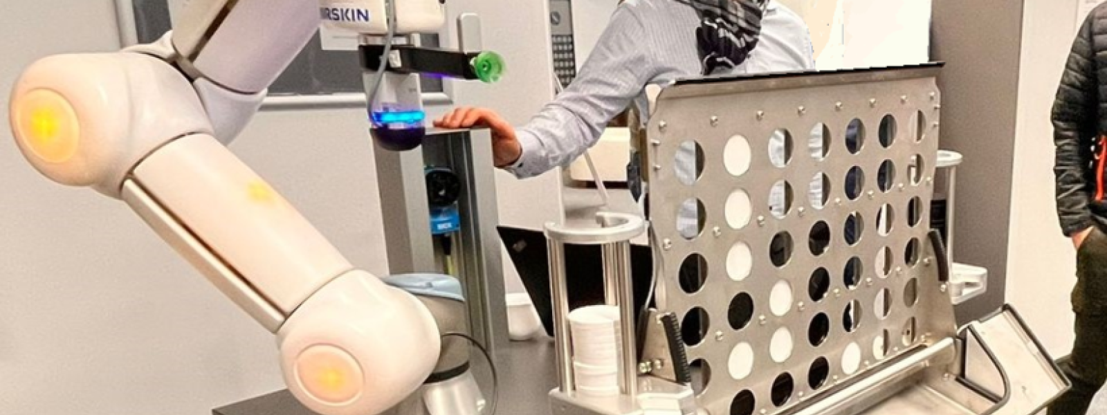 Ein Mann steht hinter einem Laboraufbau mit Roboter und lacht in die Kamera