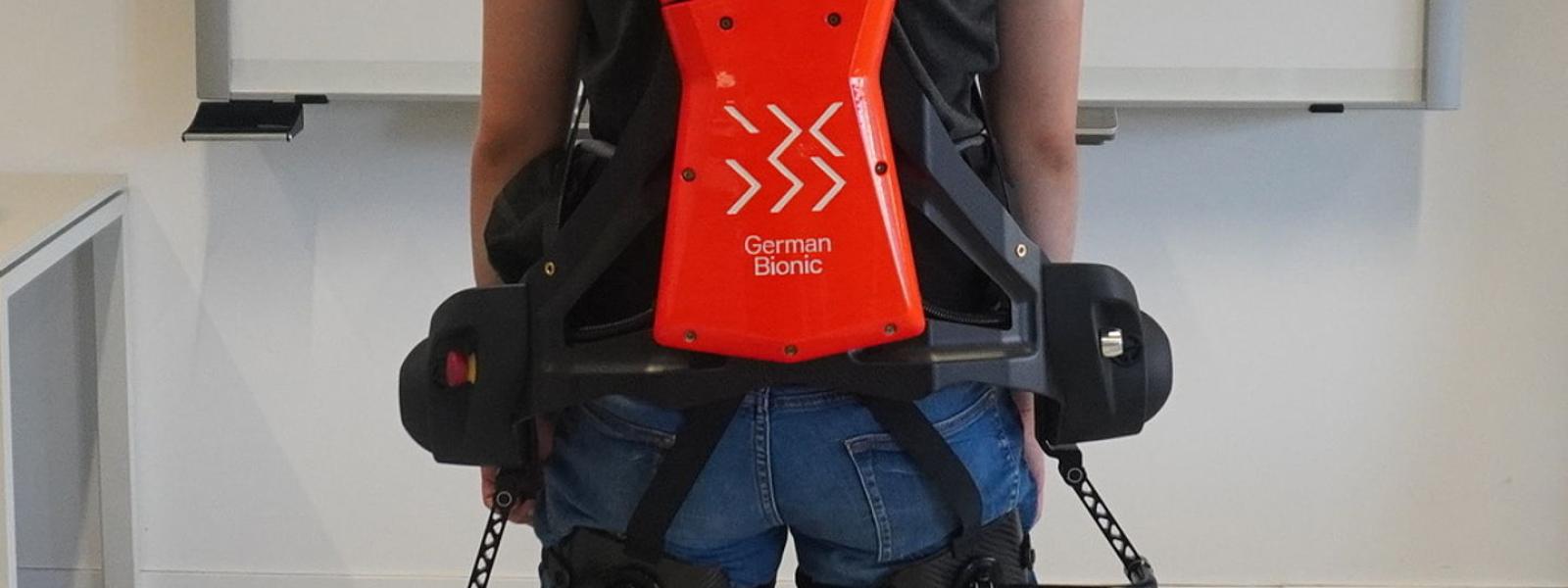 Eine Person steht mit einem angelegten Exoskelett in einem Raum vor einer weißen Tafel